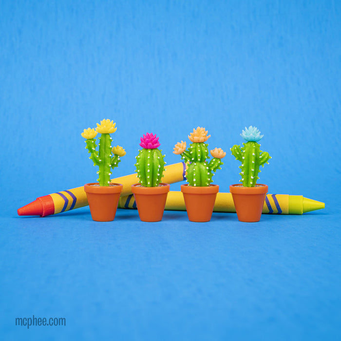 Four Itty Bitty Cactus erasers, each nestled in a pot and embellished with vibrant flowers, are displayed next to two oversized crayons on a blue background.