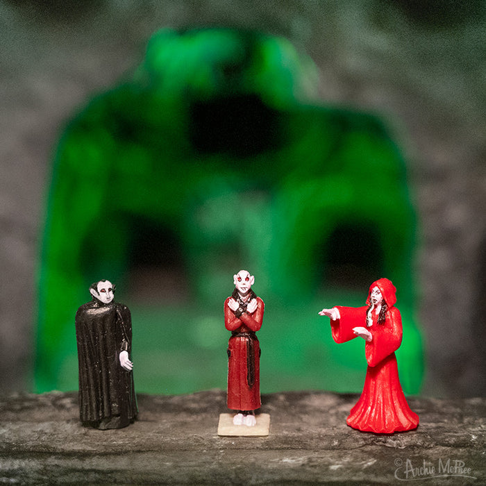 A trio of Itty Bitty Vampires is positioned against an eerie setting, showcasing a green-lit cave in the background.