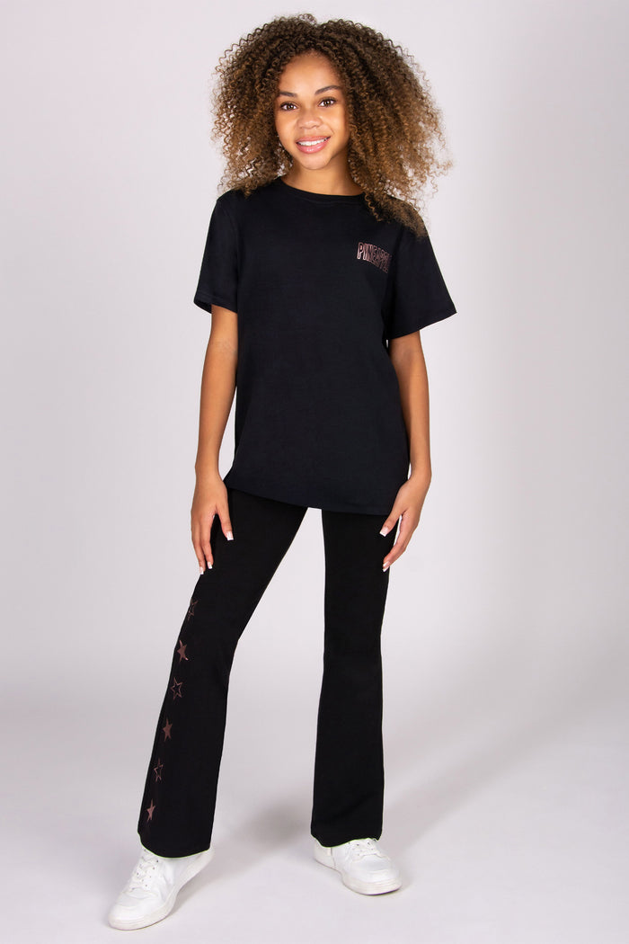 Girl wearing a black t-shirt and black flares with white shoes on a light grey background
