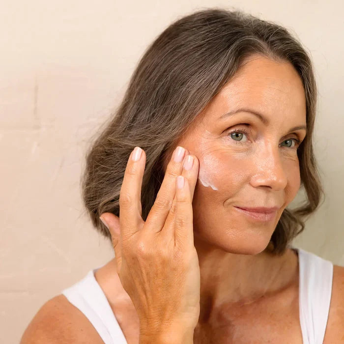 Mature woman applying cream to her face with a neutral background.