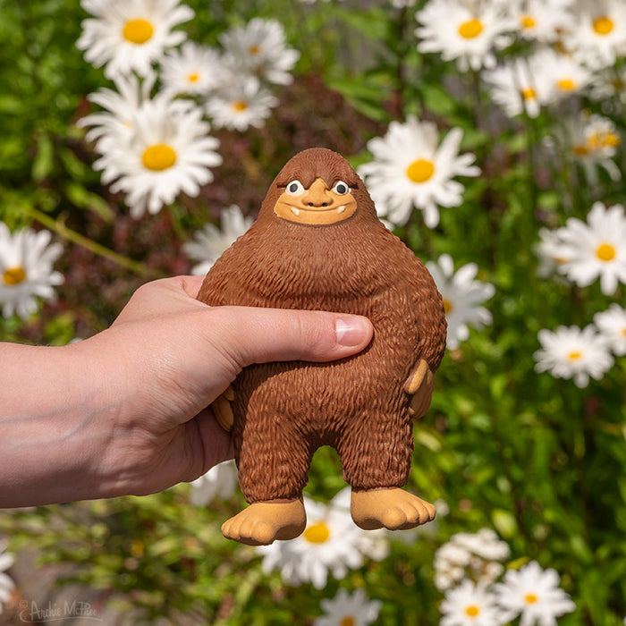 A hand holds a small, squeezable Squishy Bigfoot BFF figure against a backdrop of blooming daisies.