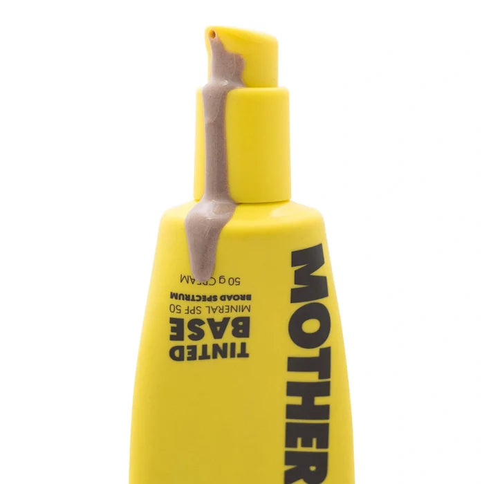 A close-up of a yellow Mother tinted base pump bottle with a beige-coloured tinted sunscreen dripping from the nozzle, on a white background.