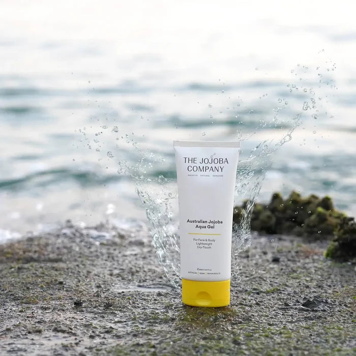 Tube of aqua gel pictured on a sandy/rocky edge next to the sea with a splash of water surrounding the product.