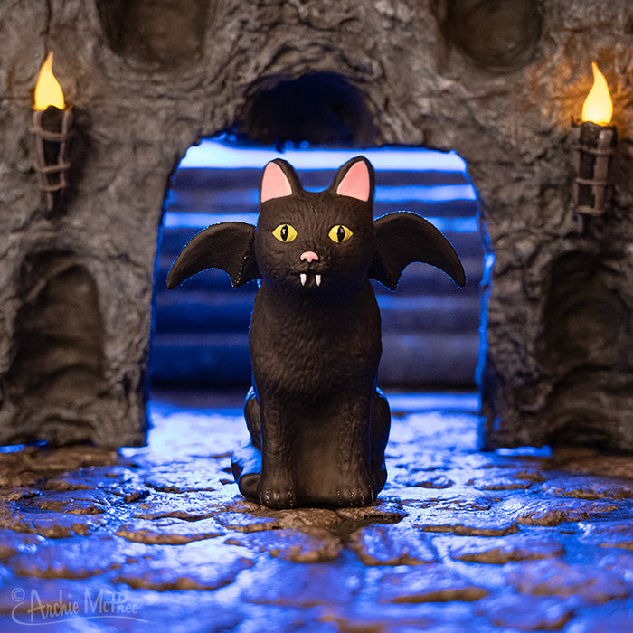 A Goth Cat Ornament with fangs sits in a dimly lit, stone archway with glowing torches.