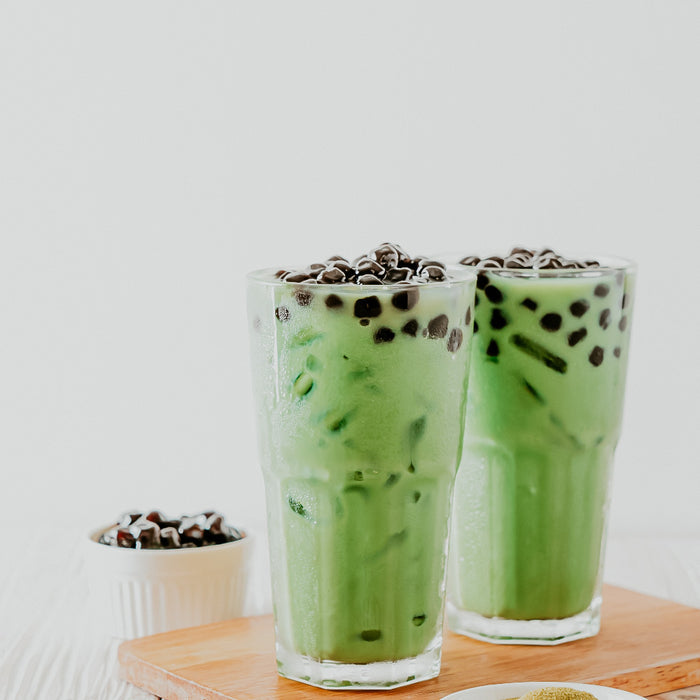 Two glasses of honeydew milk tea with boba toppings, served on a wooden board.