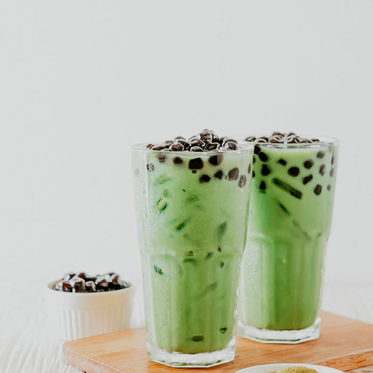 Two glasses of honeydew milk tea with boba toppings, served on a wooden board.