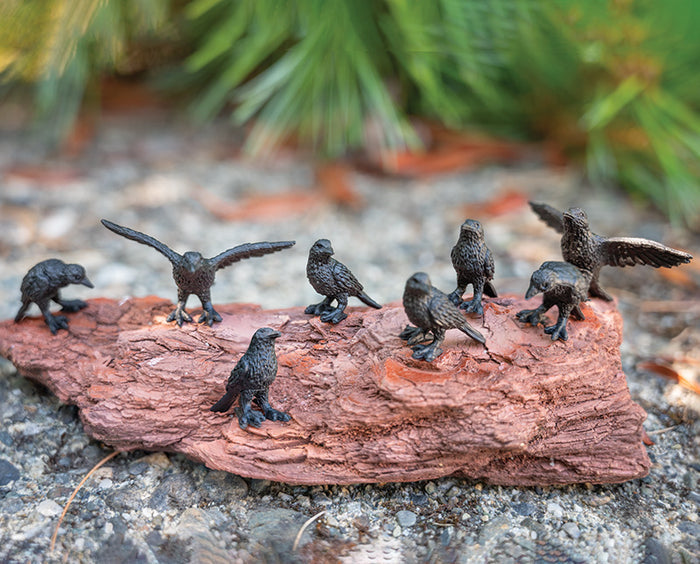 Seven Itty Bitty Crows fashioned from soft vinyl sit atop a piece of bark, surrounded by lush greenery in the background.