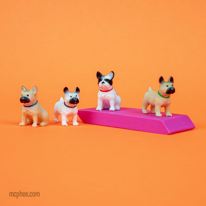 Four Itty Bitty French Bulldogs are arranged against an orange backdrop. One of these delightful figurines sits on a pink wedge, bringing a vibrant touch to the display.