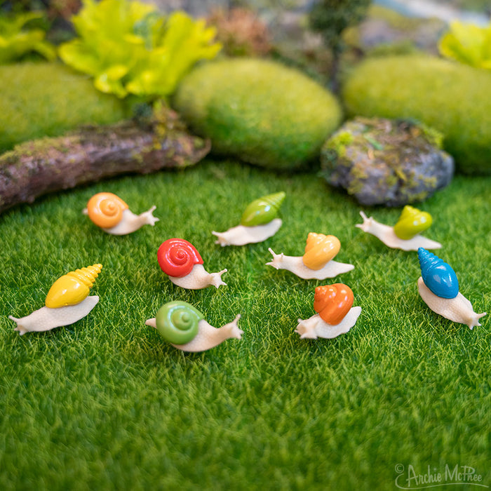 Colorful plastic Itty Bitty Snails rest on a patch of green artificial grass, with a backdrop featuring blurred plants and rocks.