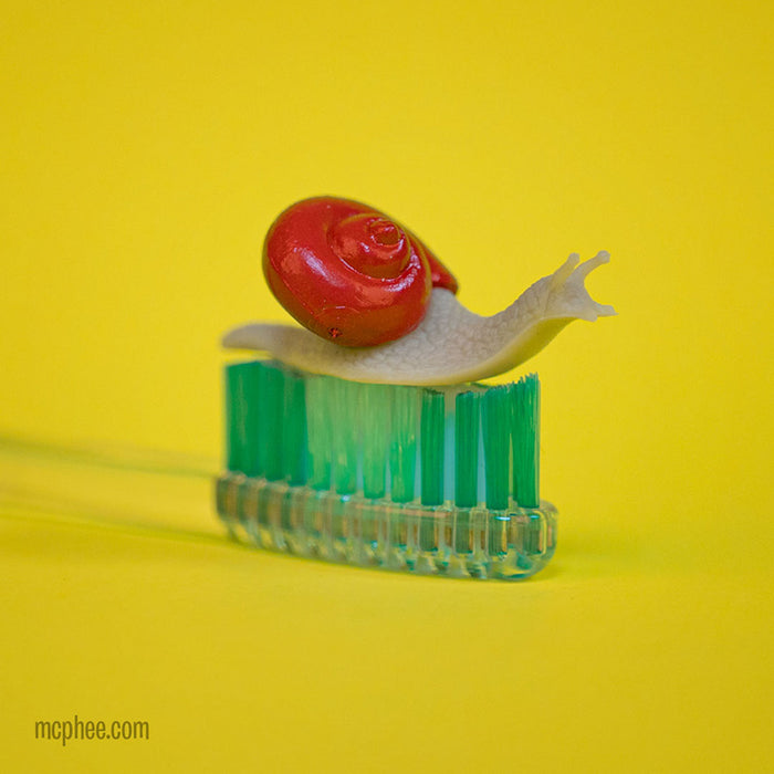 An Itty Bitty Snails miniature is perched on a toothbrush against a bright yellow background.
