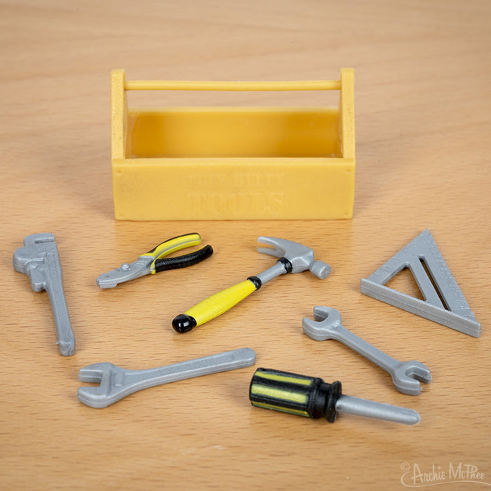 The Itty Bitty Tools set includes a yellow toolbox filled with essential compact carpentry tools, including a hammer, pliers, wrench, screwdriver, and triangle ruler on the table.
