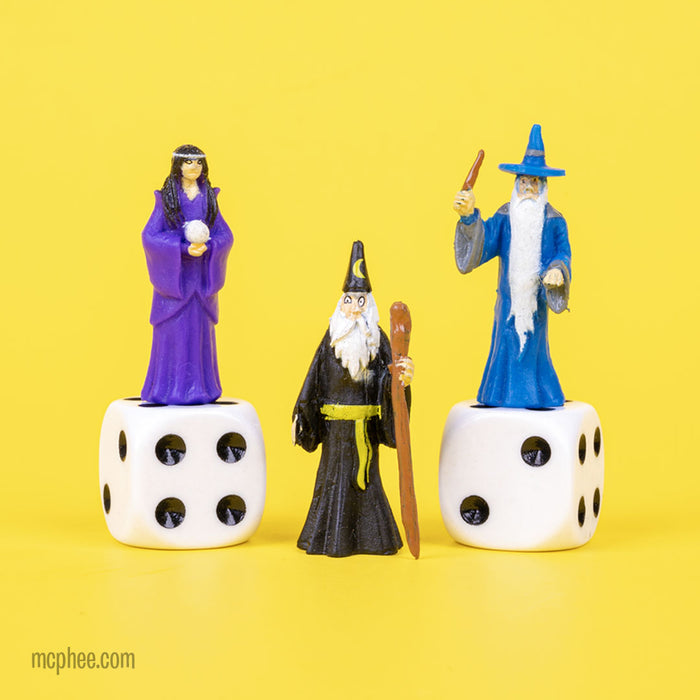 The Itty Bitty Wizards, made from soft vinyl, are arranged on dice. One is dressed in black, while the others wear robes of blue and purple, all displayed against a vibrant yellow backdrop.