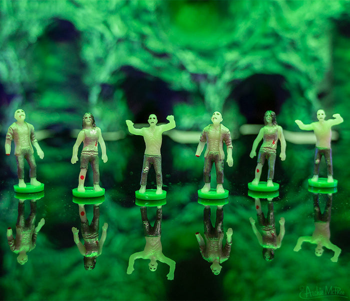 The Itty Bitty Zombies are perched on green bases, with their soft vinyl surfaces glistening on a polished surface in front of a softly focused green background.