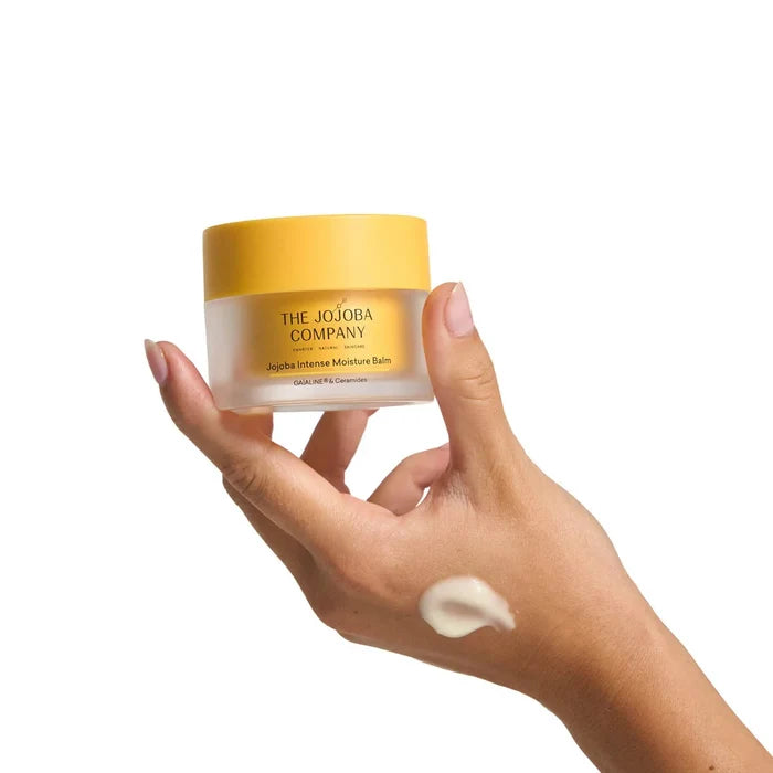Hand holding a jar of 'The Jojoba Company' balm with a visible cream smear on skin.
