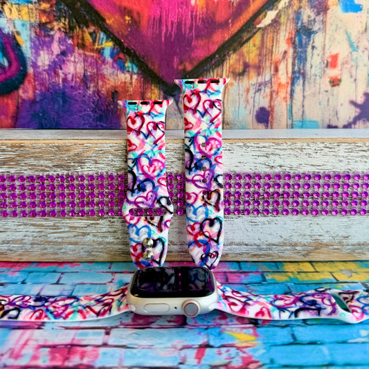 Love Out Loud silicone Apple Watch band with hand-drawn graffiti-style hearts in pink, magenta, blue, teal, red, and black on a white base, shown displayed and worn on Apple Watch against a colorful street art backdrop.