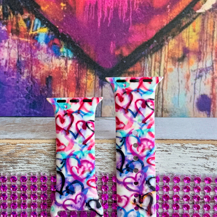 Love Out Loud silicone Apple Watch band with hand-drawn graffiti-style hearts in pink, magenta, blue, teal, red, and black on a white base, shown displayed and worn on Apple Watch against a colorful street art backdrop.