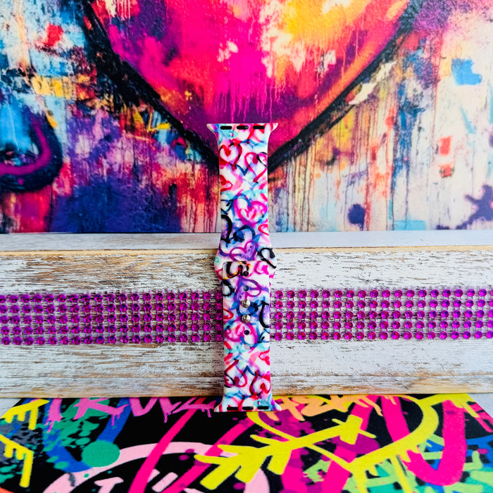 Love Out Loud silicone Apple Watch band with hand-drawn graffiti-style hearts in pink, magenta, blue, teal, red, and black on a white base, shown displayed and worn on Apple Watch against a colorful street art backdrop.