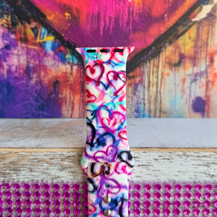 Love Out Loud silicone Apple Watch band with hand-drawn graffiti-style hearts in pink, magenta, blue, teal, red, and black on a white base, shown displayed and worn on Apple Watch against a colorful street art backdrop.
