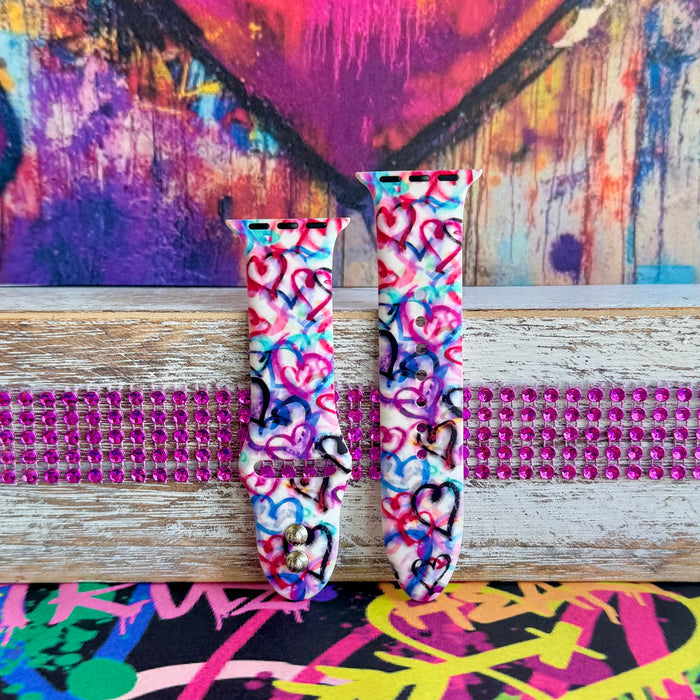 Love Out Loud silicone Apple Watch band with hand-drawn graffiti-style hearts in pink, magenta, blue, teal, red, and black on a white base, shown displayed and worn on Apple Watch against a colorful street art backdrop.