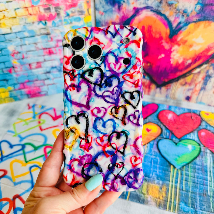 Hand holding the Love Out Loud Wavy Luxe MagSafe iPhone case, showing the graffiti-style print with layered hearts in pink, blue and purple.