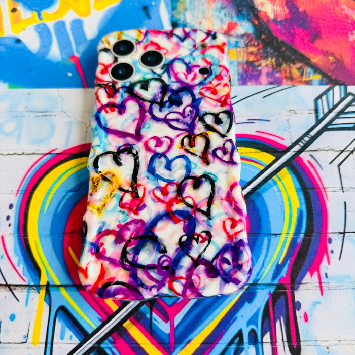 Love Out Loud Wavy Luxe iPhone case with the matching MagSafe grip ring attached, styled on a rainbow graffiti heart backdrop.
