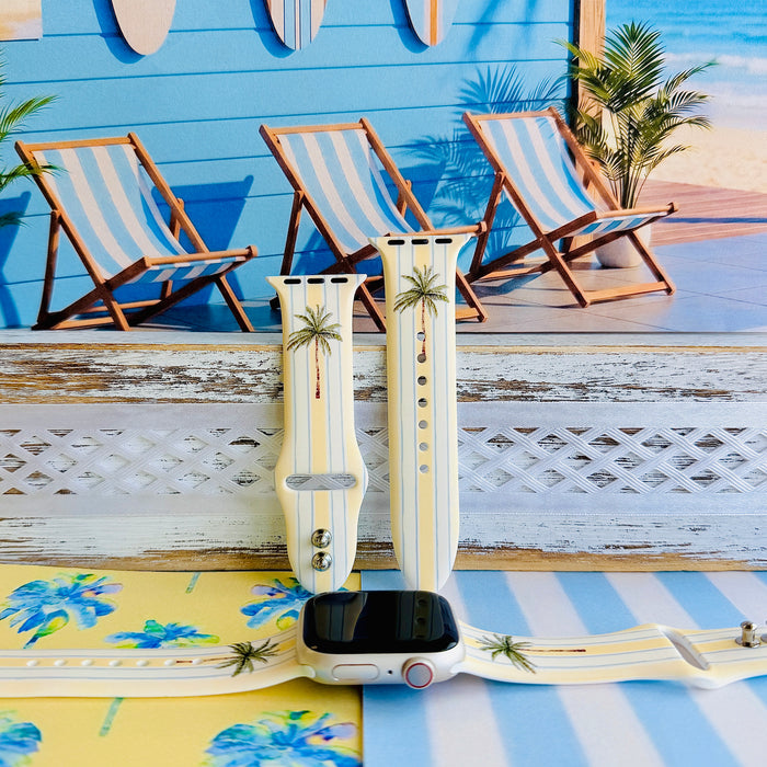 Palm Shore Club silicone Apple Watch band with hand-painted palm trees over soft blue and yellow cabana stripes on an ivory base, shown displayed and worn on Apple Watch against a coastal beach club backdrop.