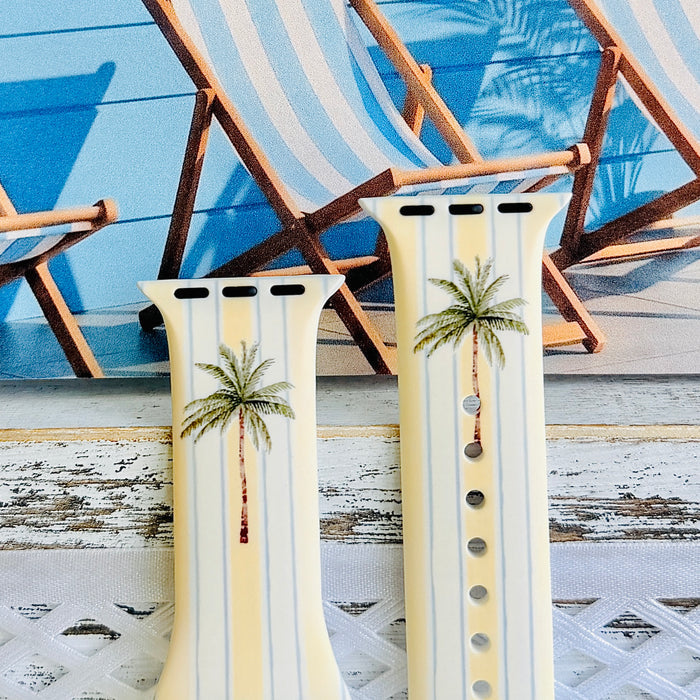 Palm Shore Club silicone Apple Watch band with hand-painted palm trees over soft blue and yellow cabana stripes on an ivory base, shown displayed and worn on Apple Watch against a coastal beach club backdrop.