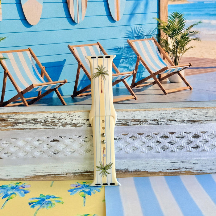 Palm Shore Club silicone Apple Watch band with hand-painted palm trees over soft blue and yellow cabana stripes on an ivory base, shown displayed and worn on Apple Watch against a coastal beach club backdrop.