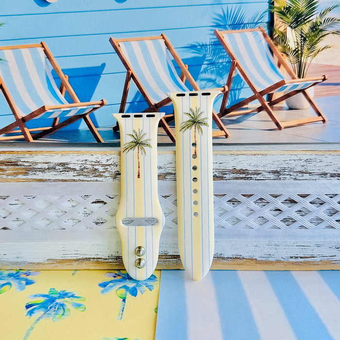 Palm Shore Club silicone Apple Watch band with hand-painted palm trees over soft blue and yellow cabana stripes on an ivory base, shown displayed and worn on Apple Watch against a coastal beach club backdrop.