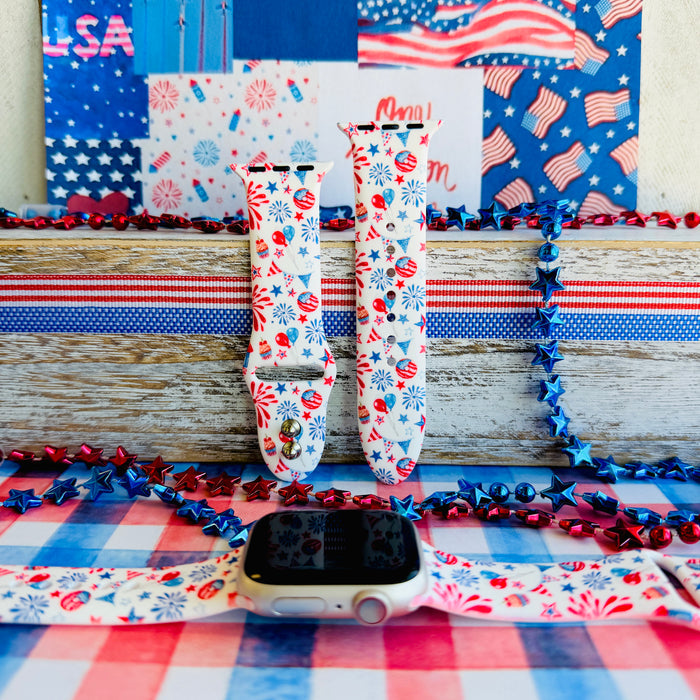 Party In The USA silicone Apple Watch band with fireworks, balloons, cupcakes, flag bunting, and stars in red, white, and blue on a cream base, shown displayed and worn on Apple Watch against a patriotic 4th of July backdrop.