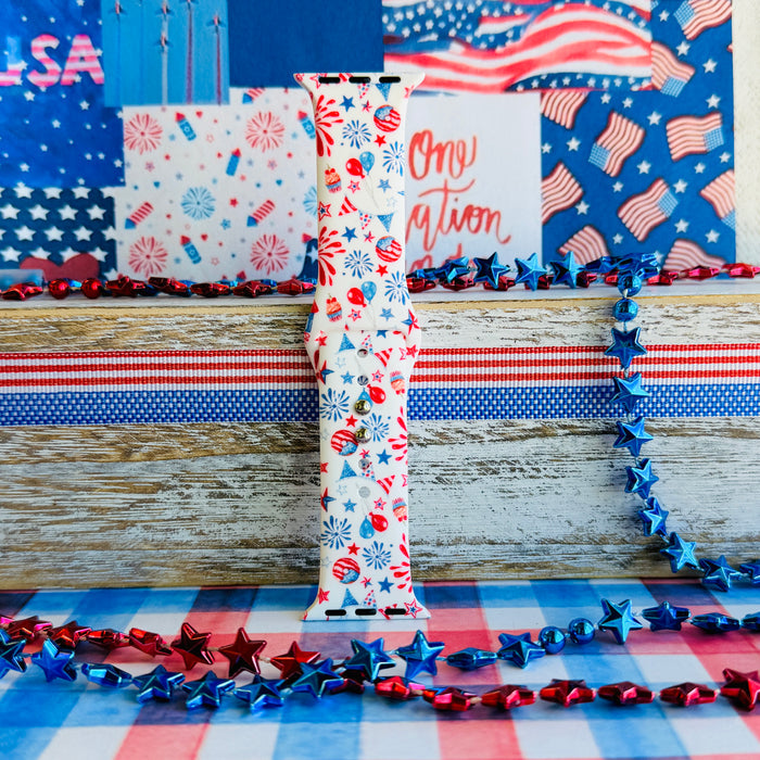 Party In The USA silicone Apple Watch band with fireworks, balloons, cupcakes, flag bunting, and stars in red, white, and blue on a cream base, shown displayed and worn on Apple Watch against a patriotic 4th of July backdrop.