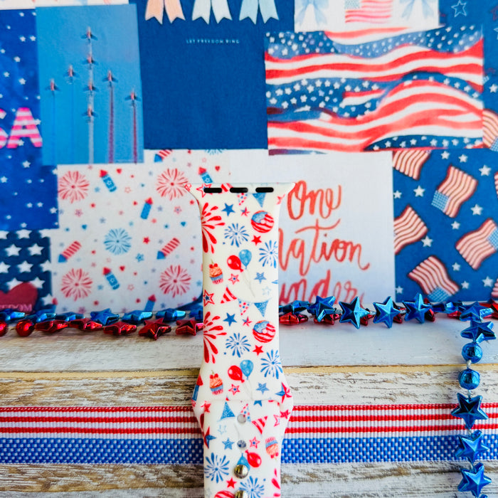 Party In The USA silicone Apple Watch band with fireworks, balloons, cupcakes, flag bunting, and stars in red, white, and blue on a cream base, shown displayed and worn on Apple Watch against a patriotic 4th of July backdrop.