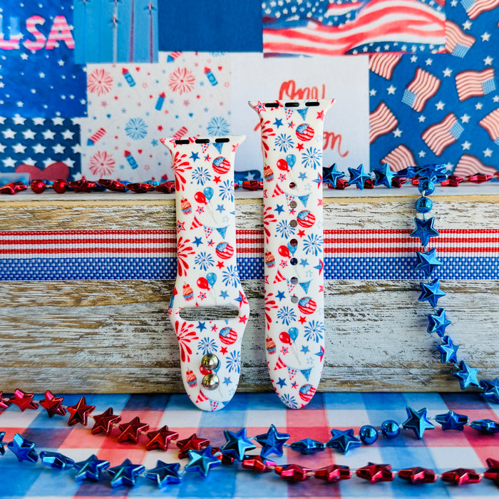 Party In The USA silicone Apple Watch band with fireworks, balloons, cupcakes, flag bunting, and stars in red, white, and blue on a cream base, shown displayed and worn on Apple Watch against a patriotic 4th of July backdrop.