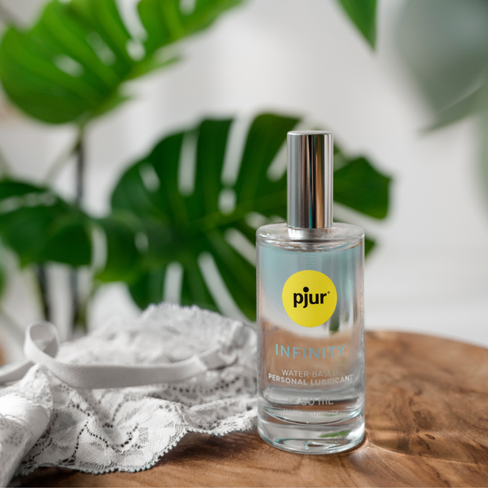 A close-up, stylized photo of a clear glass bottle of pjur Infinity Water-Based Personal Lubricant, 50ml, with a silver cap. The bottle is placed on a wooden surface next to a piece of white lace lingerie.