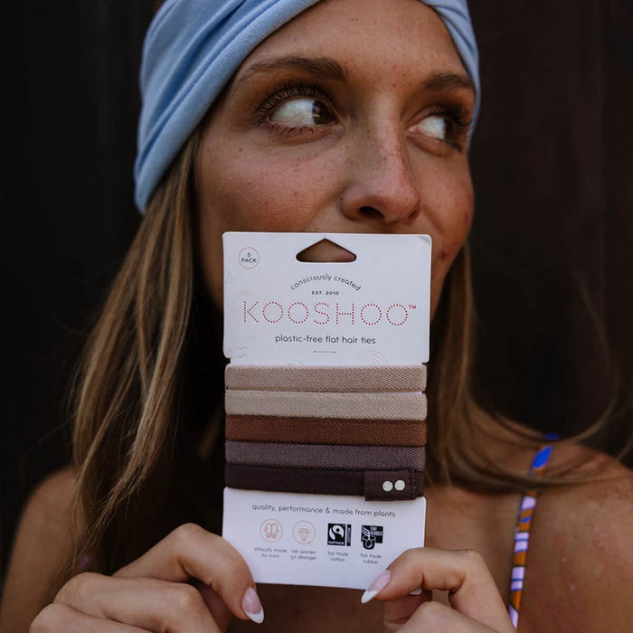 Woman holding a package of Kooshoo Plastic-Free hair ties against her face with a dark background.