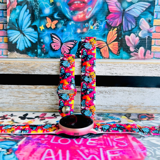 Rebel Wings silicone Samsung Galaxy Watch band with graffiti-style butterflies in blue, orange, yellow, and white over a hot pink and magenta paint-splatter background, shown displayed and worn against a colorful street art butterfly backdrop.