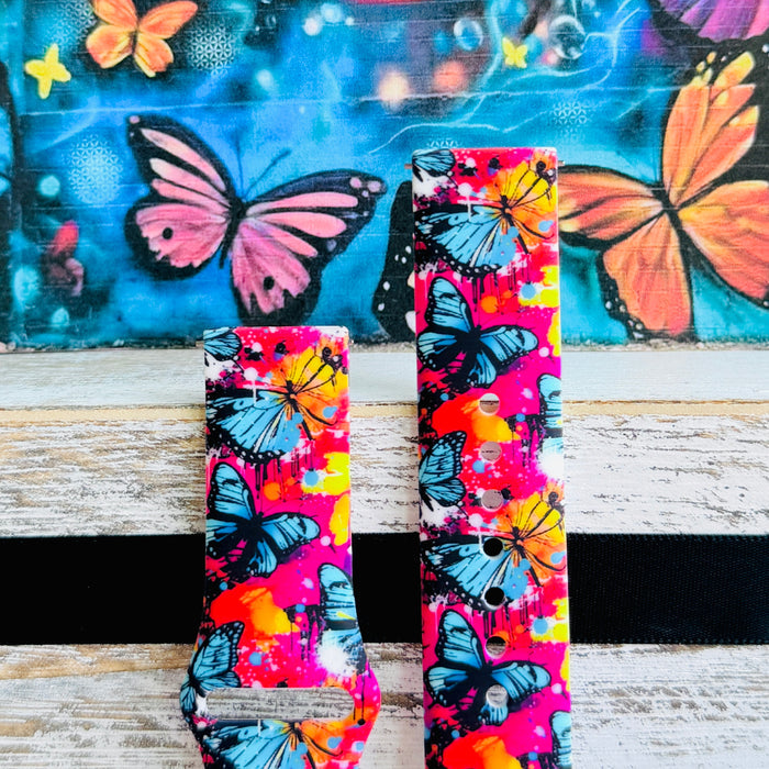Rebel Wings silicone Samsung Galaxy Watch band with graffiti-style butterflies in blue, orange, yellow, and white over a hot pink and magenta paint-splatter background, shown displayed and worn against a colorful street art butterfly backdrop.