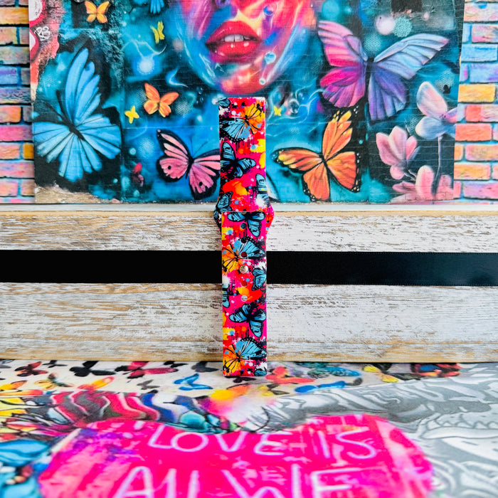 Rebel Wings silicone Samsung Galaxy Watch band with graffiti-style butterflies in blue, orange, yellow, and white over a hot pink and magenta paint-splatter background, shown displayed and worn against a colorful street art butterfly backdrop.
