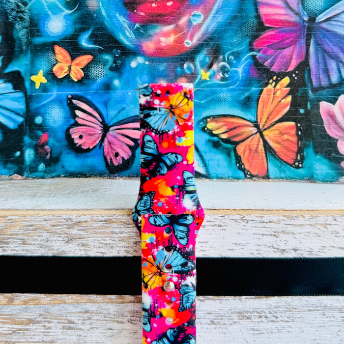 Rebel Wings silicone Samsung Galaxy Watch band with graffiti-style butterflies in blue, orange, yellow, and white over a hot pink and magenta paint-splatter background, shown displayed and worn against a colorful street art butterfly backdrop.