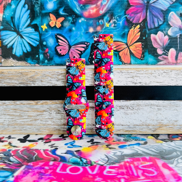Rebel Wings silicone Samsung Galaxy Watch band with graffiti-style butterflies in blue, orange, yellow, and white over a hot pink and magenta paint-splatter background, shown displayed and worn against a colorful street art butterfly backdrop.