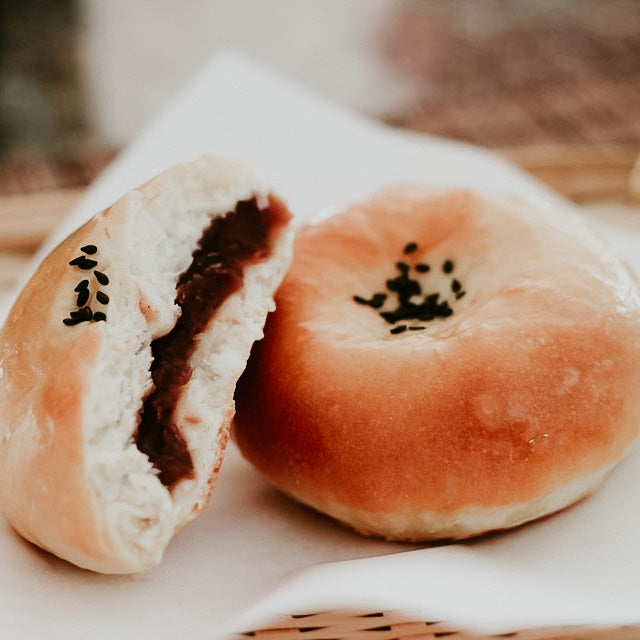 A photo of a red bean bun, showing the bread roll with a visible filling and black sesame seeds on top.