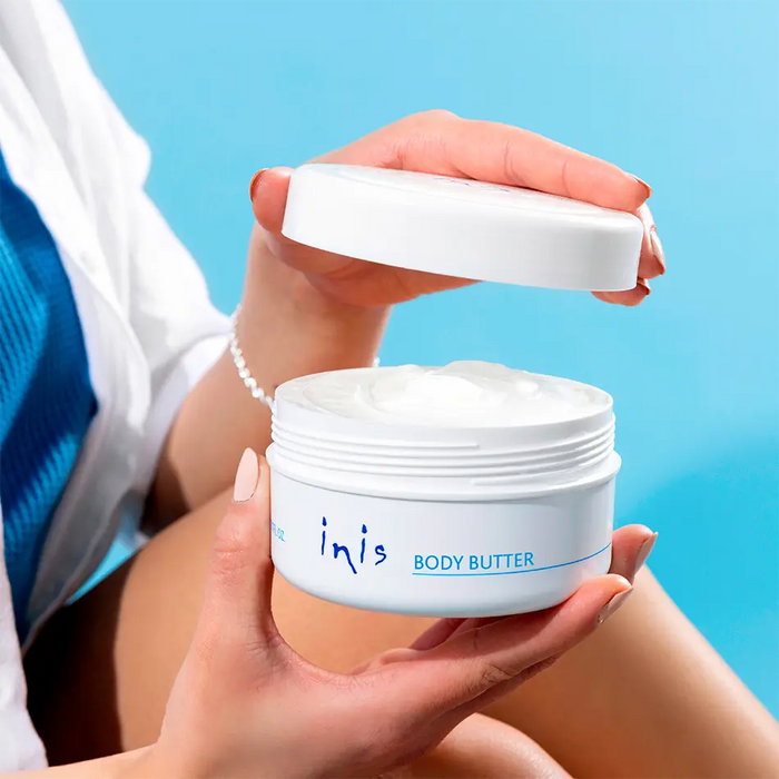 A person holds an open white jar of inis BODY BUTTER, showing the white cream inside, against a bright blue background.