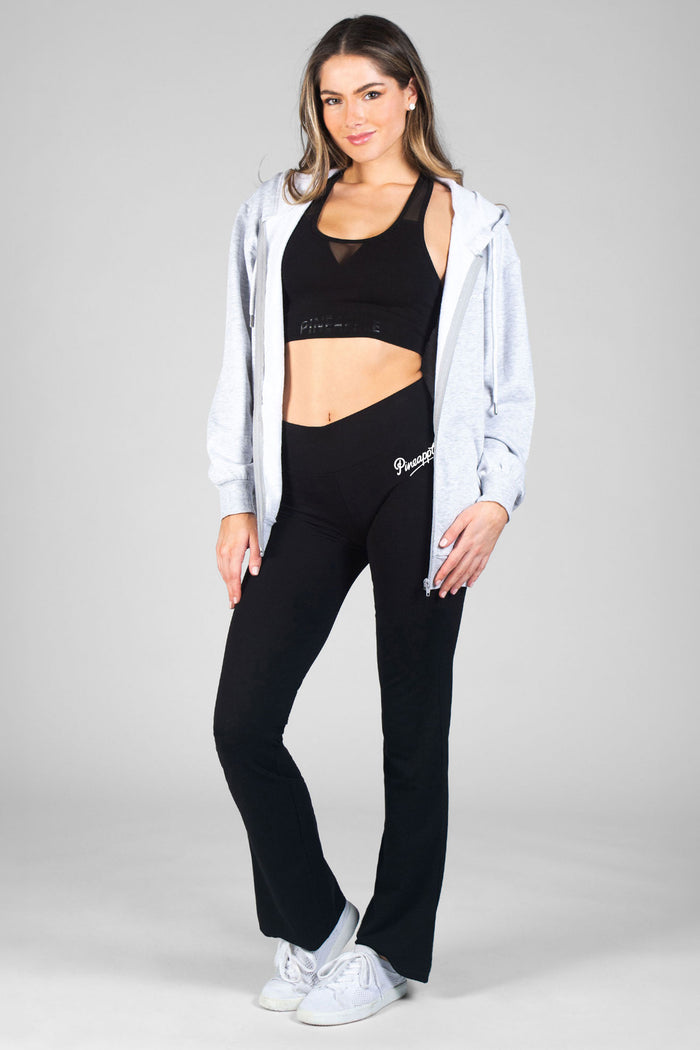 Woman wearing black flared yoga pants with black crop top and grey zip hoodie against a grey background.