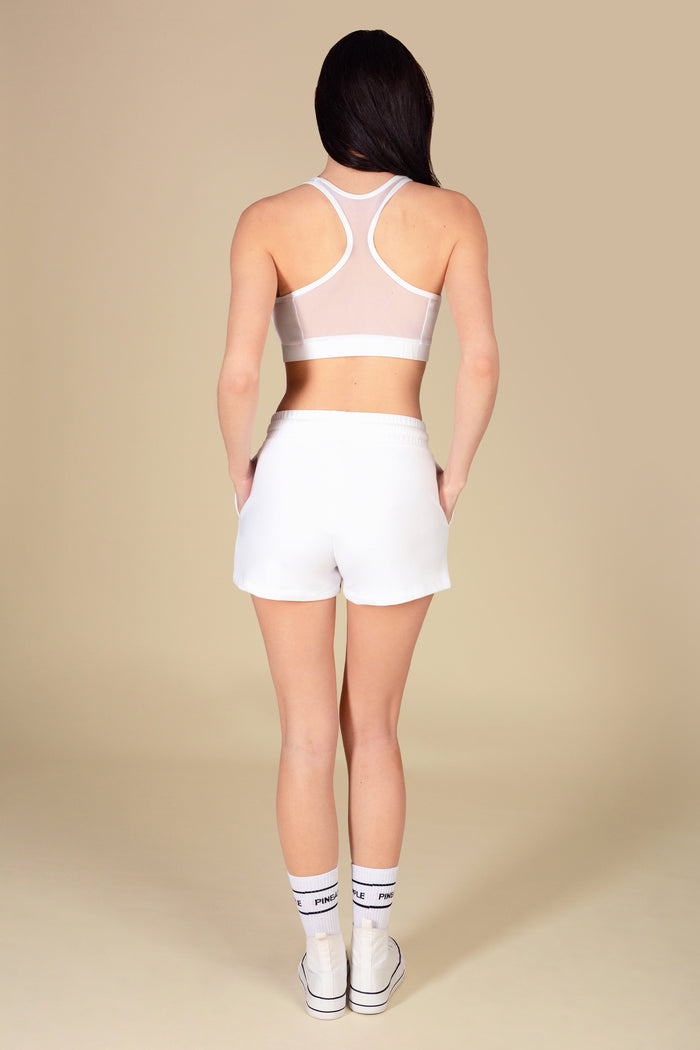 Woman wearing an athletic outfit with white mesh bra top and white pocket retro shorts on a beige background.