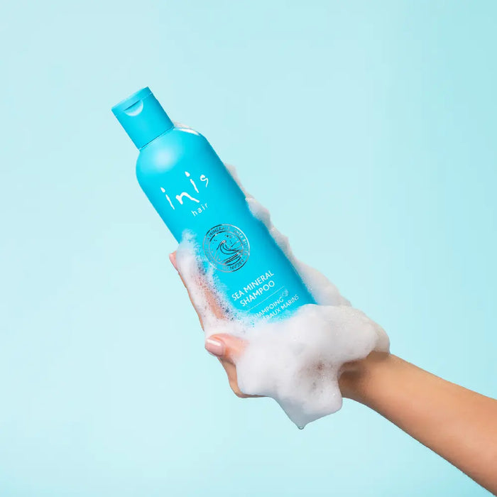 A hand holding a turquoise blue Inis Sea Mineral Shampoo bottle covered in white foam, against a solid light blue background.