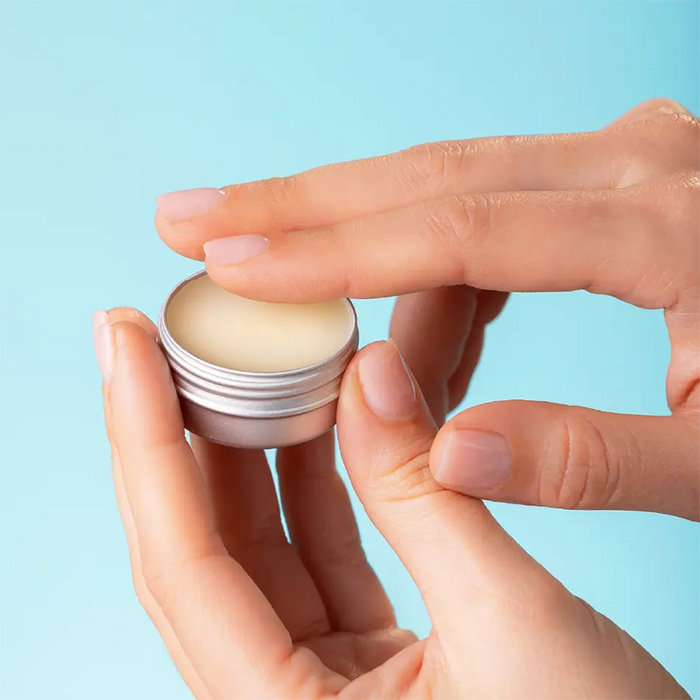 A close-up of hands applying a lip balm from a small silver metal tin against a light blue background.