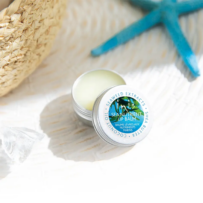 An open round tin of lip balm sits on a white textured surface with a straw hat, blue starfish. The lid label reads 'inis Sea Nutriments Lip Balm' with coconut oil, seaweed extracts, and sea butter text.
