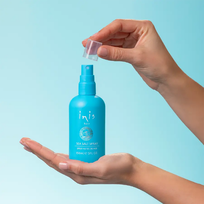 A person's hands holding a turquoise blue Inis Sea Salt Spray bottle, with one hand unscrewing the clear cap while the other supports the bottle against a solid light blue background.