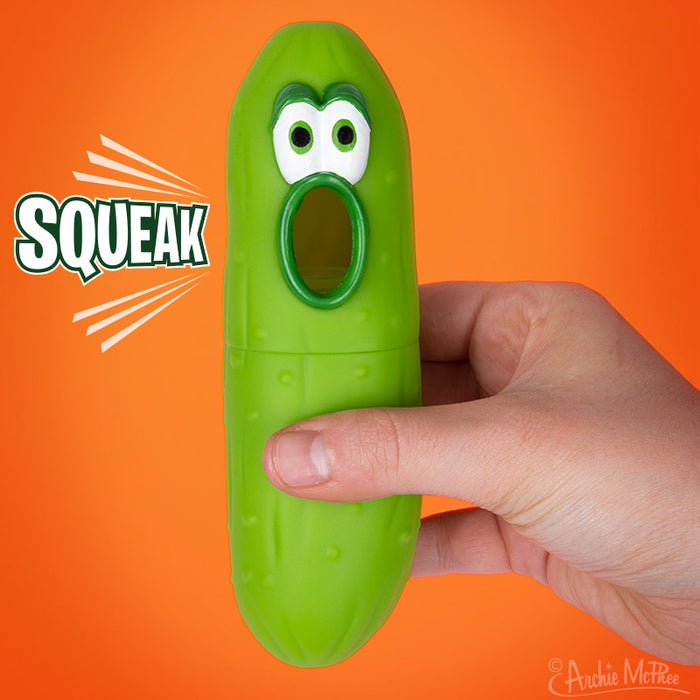 A hand holds a squeaky green Squeak Pickle toy with cartoon eyes and an open mouth, set against an orange background. This playful item brings to mind the whimsical designs by Archie McPhee.