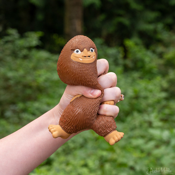 A hand squeezes a smiling Bigfoot toy made of soft rubber against a blurred green background, capturing a playful moment with the Squishy Bigfoot BFF.